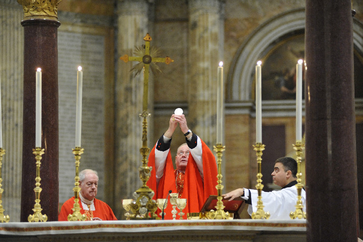 Cardinal Dolan elevates the Eucharist during the final Mass of the pilgrimage to Rome at the Basilica of St. Paul Outside the Walls Feb. 20. With him at the altar is Cardinal Egan, Archbishop Emeritus of New York and a fellow member of the College of Cardinals. Overall view at left shows concelebrating bishops and pilgrims.