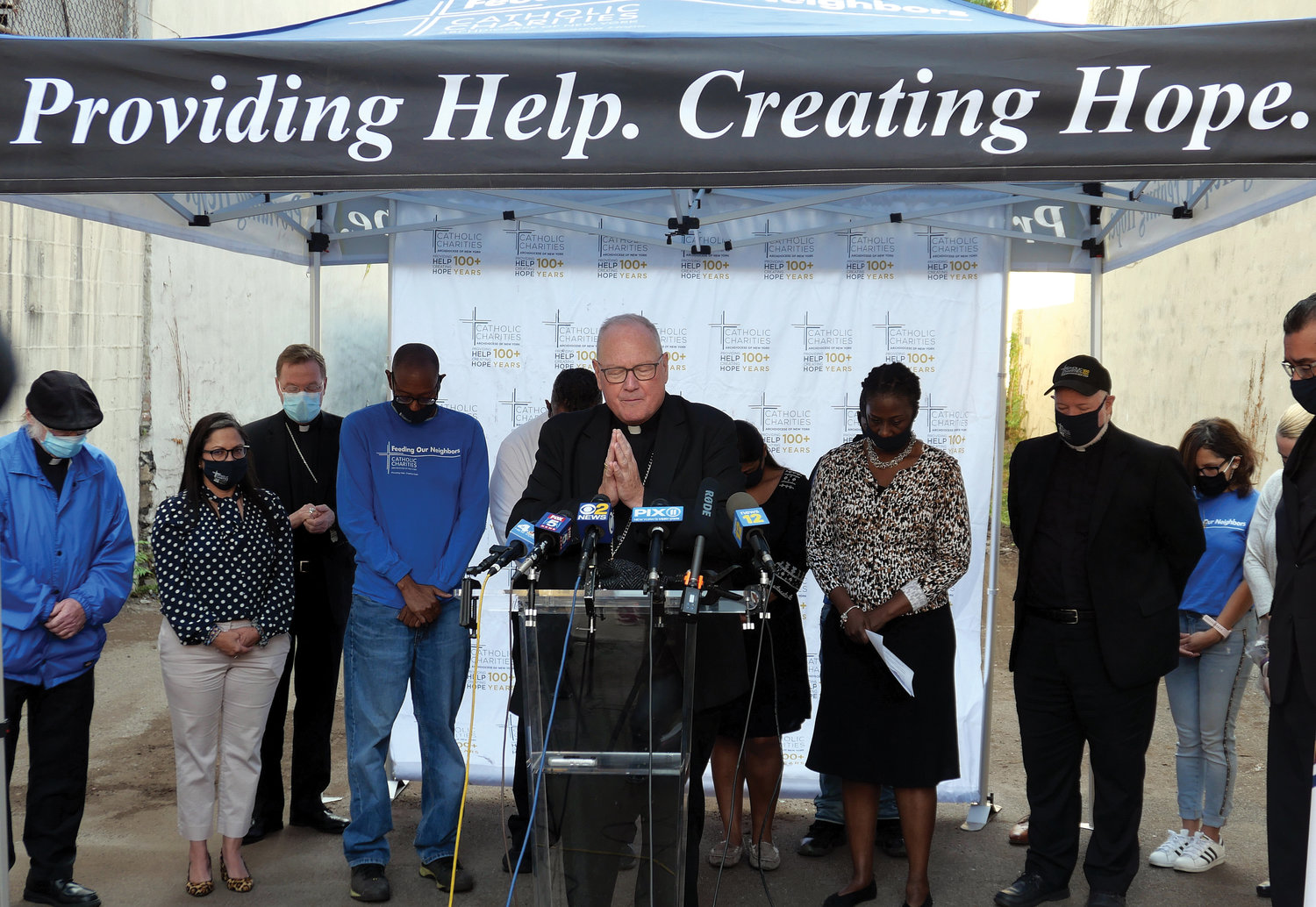 Cardinal Dolan offers a blessing at the start of a Sept. 23 Catholic Charities food distribution gathering at a Catholic Charities site on East 152nd Street in the Bronx. Among the speakers was Msgr. Kevin Sullivan, executive director of archdiocesan Catholic Charities, seen at right with cap.