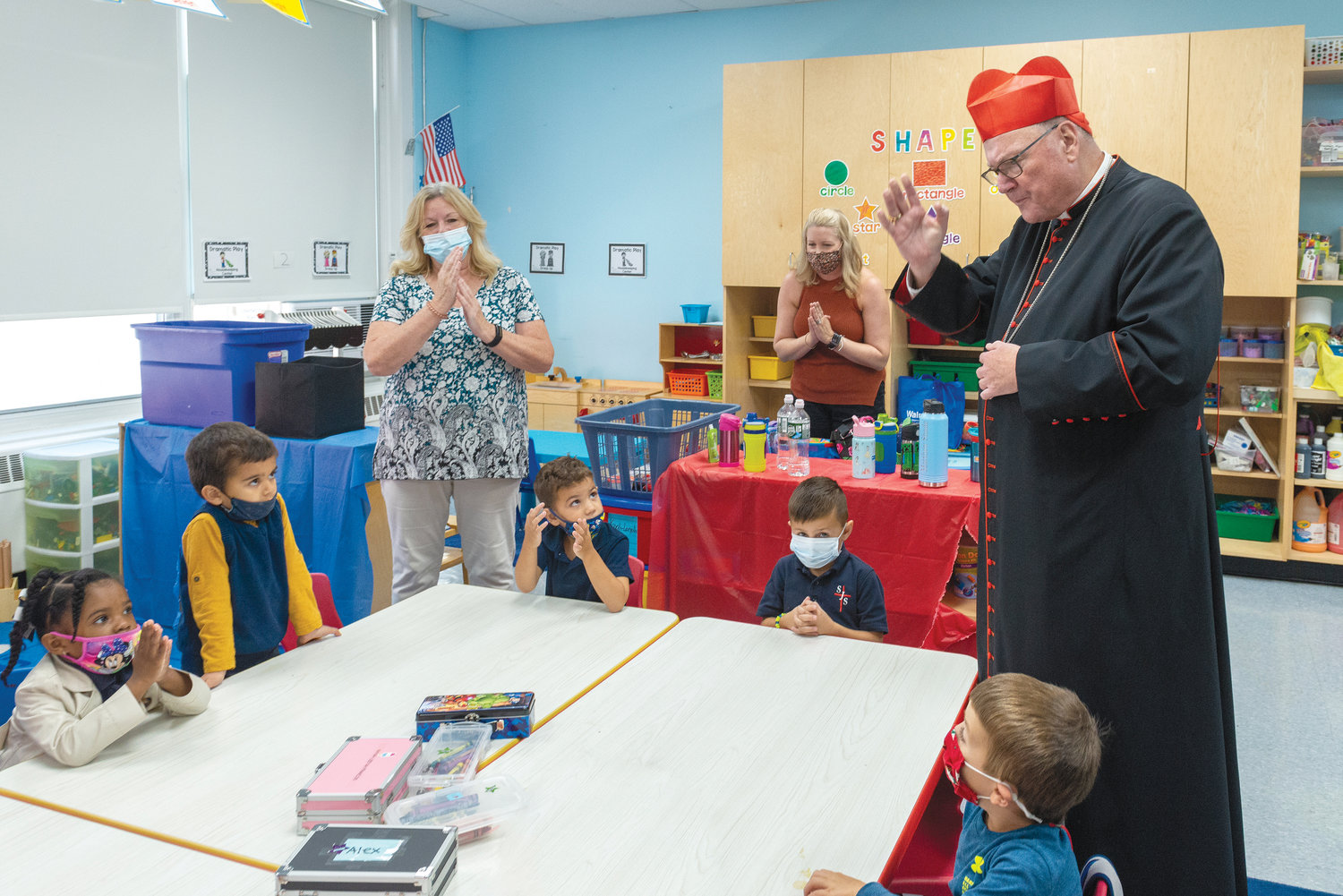 The cardinal visits with young students and staff in a classroom.